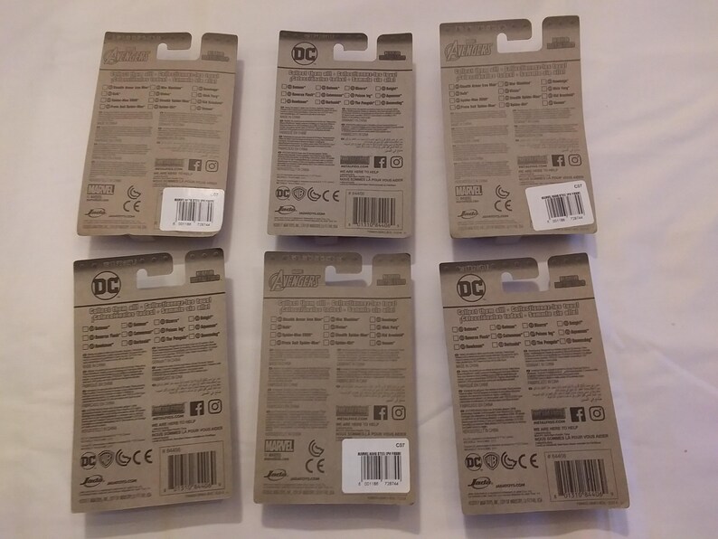 May include: Six toy action figure packages with brown cardboard backing. The packages feature the logos for DC and Marvel comics, with text and barcodes visible. The packages are arranged on a white surface.