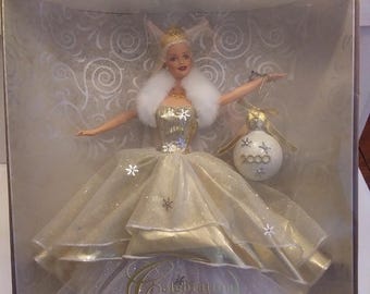May include: A boxed Barbie doll wearing a white and gold gown with a white fur collar. The doll is holding a gold and white ornament that says "2000". The box has a gold border and says "Celebration Barbie" on the bottom.