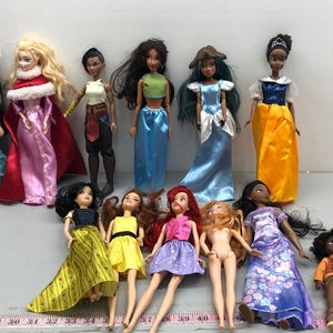 May include: A collection of fashion dolls, featuring various characters in colorful gowns and outfits. The dolls have detailed facial features and hairstyles, with a variety of skin tones and clothing styles. Some dolls are standing, while others are lying down.