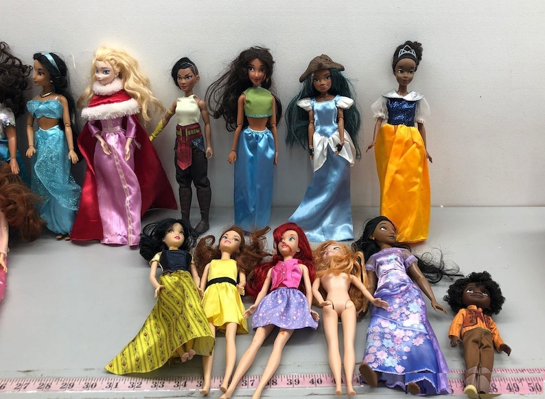 May include: A collection of fashion dolls, featuring various characters in colorful gowns and outfits. The dolls have detailed facial features and hairstyles, with a variety of skin tones and clothing styles. Some dolls are standing, while others are lying down.