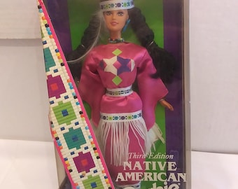 May include: A Native American Barbie doll in a box. The doll wears a pink dress with a white fringe skirt and a beaded headband. The box is pink and has the words "Special Edition" and "Dolls of the World Collection."