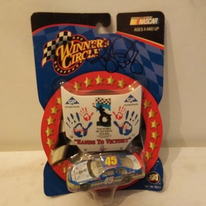 Op de afbeelding: Een blauwe en witte NASCAR-modelwagen met nummer 45 aan de zijkant. De auto is verpakt in een plastic blisterverpakking met een rode en gouden achtergrond. De verpakking heeft de tekst "Winner's Circle" en "Hands to Victory".