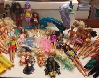 May include: A collection of dolls, including Barbie dolls, Disney princesses, and other characters. The dolls are arranged on a white surface with a blue and purple dragon toy in the center.