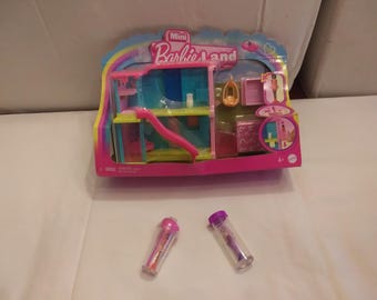 May include: A Mini Barbie Land playset in a pink and blue box, featuring a slide, swing, and accessories. Two small, clear tubes with pink and purple tops are in front of the playset.
