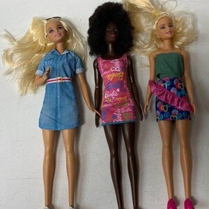May include: Three Barbie dolls wearing different outfits. The doll on the left is wearing a blue denim dress with a white belt. The doll in the middle is wearing a pink dress with a floral pattern and the text "Barbie" repeated on the dress. The doll on the right is wearing a green tank top and a pink and blue skirt with a ruffle.
