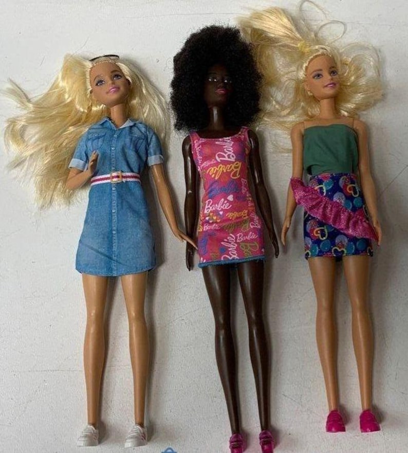 May include: Three Barbie dolls wearing different outfits. The doll on the left is wearing a blue denim dress with a white belt. The doll in the middle is wearing a pink dress with a floral pattern and the text "Barbie" repeated on the dress. The doll on the right is wearing a green tank top and a pink and blue skirt with a ruffle.