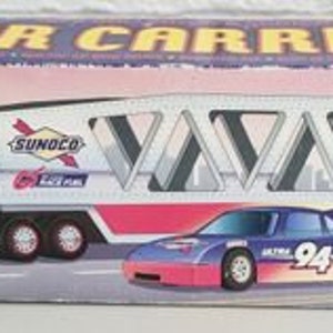 Puede incluir: Un transportador de coches de juguete azul y blanco con las palabras "Sunoco Ultra 94" en el lateral. El transportador lleva un coche de carreras rojo y blanco con el número "94" en el lateral. La caja tiene las palabras "Car Carrier" y "Sixth of a Series" en ella.