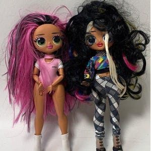May include: Two fashion dolls with long, colorful hair. One doll has pink and black hair and is wearing a pink and white outfit with the word "OMG" on it. The other doll has black and white hair and is wearing a black and white checkered outfit with a rainbow shirt.