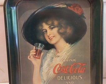 May include: A vintage Coca-Cola metal tray featuring a woman in a large hat holding a glass of Coca-Cola. The tray has a dark green border and the text "Drink Coca-Cola Delicious and Refreshing".