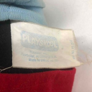 Puede incluir: Una etiqueta blanca con texto azul que dice "PLAYSKOOL" y "REG. NO. PA 228". La etiqueta tambi&eacute;n incluye la direcci&oacute;n de la empresa y el pa&iacute;s de origen. La etiqueta est&aacute; unida a una tela roja y negra.