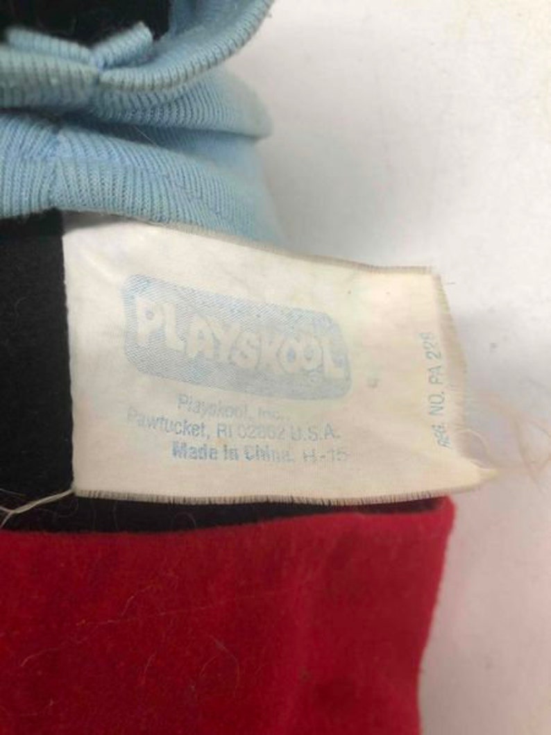 Puede incluir: Una etiqueta blanca con texto azul que dice "PLAYSKOOL" y "REG. NO. PA 228". La etiqueta tambi&eacute;n incluye la direcci&oacute;n de la empresa y el pa&iacute;s de origen. La etiqueta est&aacute; unida a una tela roja y negra.
