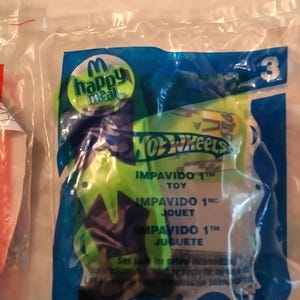 Może przedstawiać: Niebieskie i zielone opakowanie zabawki Happy Meal z autkiem Hot Wheels. Opakowanie zawiera tekst "Impavido 1" w języku angielskim, francuskim i hiszpańskim.