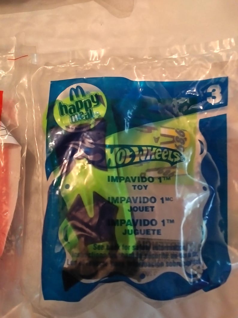 Może przedstawiać: Niebieskie i zielone opakowanie zabawki Happy Meal z autkiem Hot Wheels. Opakowanie zawiera tekst "Impavido 1" w języku angielskim, francuskim i hiszpańskim.