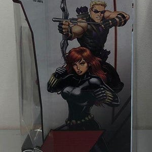 May include: A box for a Marvel Avengers toy featuring Black Widow and Hawkeye. The box is red, white and black with a picture of the characters on the front. The text "MARVEL AVENGERS" is printed in white on a red background. The box also includes instructions for use and a list of required batteries.