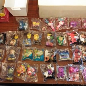 Puede incluir: Una colecci&oacute;n de 24 juguetes empaquetados individualmente, con personajes de programas de televisi&oacute;n y pel&iacute;culas infantiles populares. Los juguetes se muestran en una superficie de madera e incluyen una variedad de colores y dise&ntilde;os.