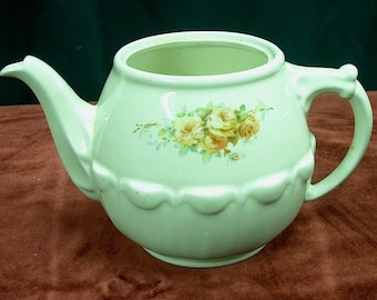 May include: A pale green teapot with a floral design of yellow roses and green leaves. The teapot has a scalloped edge and a rounded body.