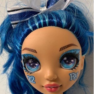 May include: A blue haired doll with blue and white glitter eye shadow and a blue and white bow in her hair. She is wearing a blue and white cheerleading uniform with the word "RAINBOW" on the front. The doll has a number "22" and the letter "RY" painted on her face.