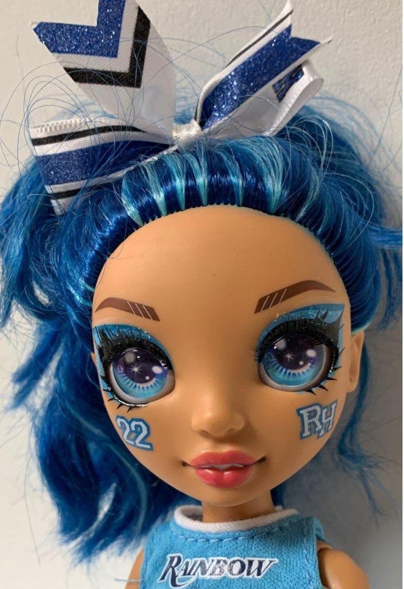 May include: A blue haired doll with blue and white glitter eye shadow and a blue and white bow in her hair. She is wearing a blue and white cheerleading uniform with the word "RAINBOW" on the front. The doll has a number "22" and the letter "RY" painted on her face.