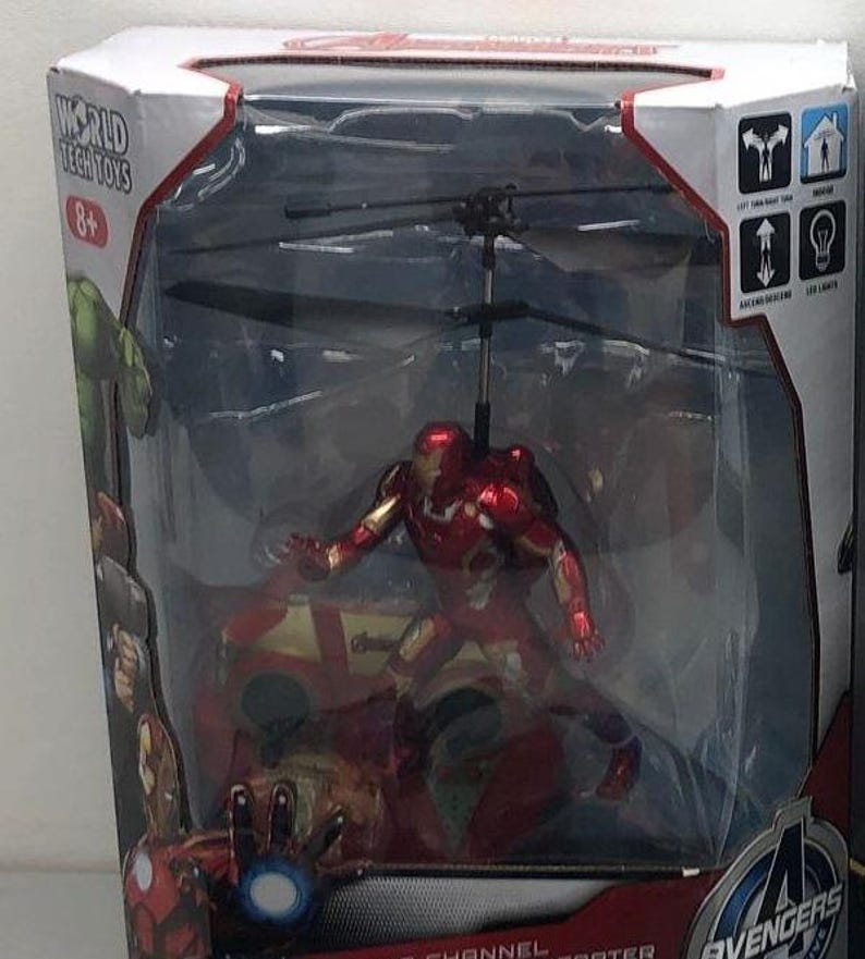May include: A red and gold Iron Man action figure in a helicopter toy. The helicopter has a clear plastic casing and a black rotor. The box has the words "Avengers" and "World Tech Toys" on it.