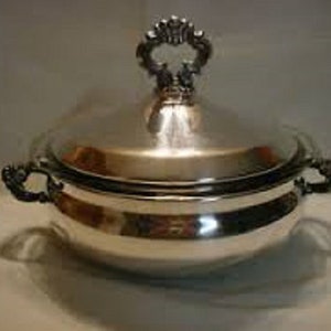 Könnte beinhalten: Eine silberne Servierplatte mit Deckel und verziertem Griff. Die Platte ist rund und hat eine glatte, polierte Oberfläche.