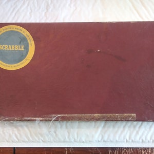 May include: A red box with a yellow circle on the front. The circle has the word "SCRABBLE" in white letters and the text "MANUFACTURED BY SELCHOW & RIGHTER CO. NEW YORK, N.Y." in black letters.