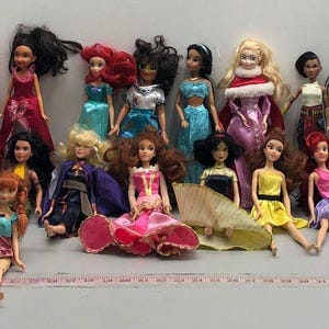 May include: A collection of dolls, each dressed in unique outfits. The dolls have various skin tones and hairstyles, with a range of colorful dresses and accessories. Some dolls are standing, while others are seated.