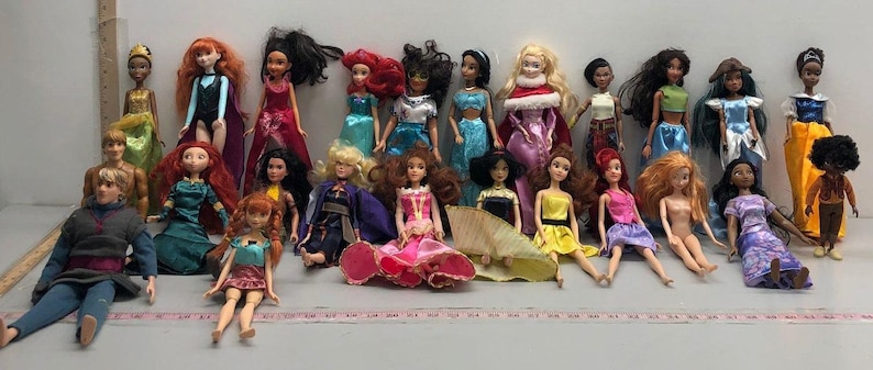 May include: A collection of dolls, each dressed in unique outfits. The dolls have various skin tones and hairstyles, with a range of colorful dresses and accessories. Some dolls are standing, while others are seated.