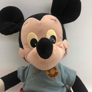 Puede incluir: Un juguete de peluche vintage de Mickey Mouse que lleva una camisa azul con un logotipo de "Mickey Mouse" rojo y blanco. El juguete tiene orejas negras, una nariz negra y grandes ojos marrones.
