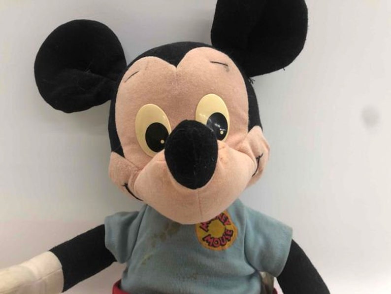 Puede incluir: Un juguete de peluche vintage de Mickey Mouse que lleva una camisa azul con un logotipo de "Mickey Mouse" rojo y blanco. El juguete tiene orejas negras, una nariz negra y grandes ojos marrones.