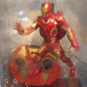 May include: A red and gold remote control toy of Iron Man flying with a red and gold remote control. The remote control has the Avengers logo on it.