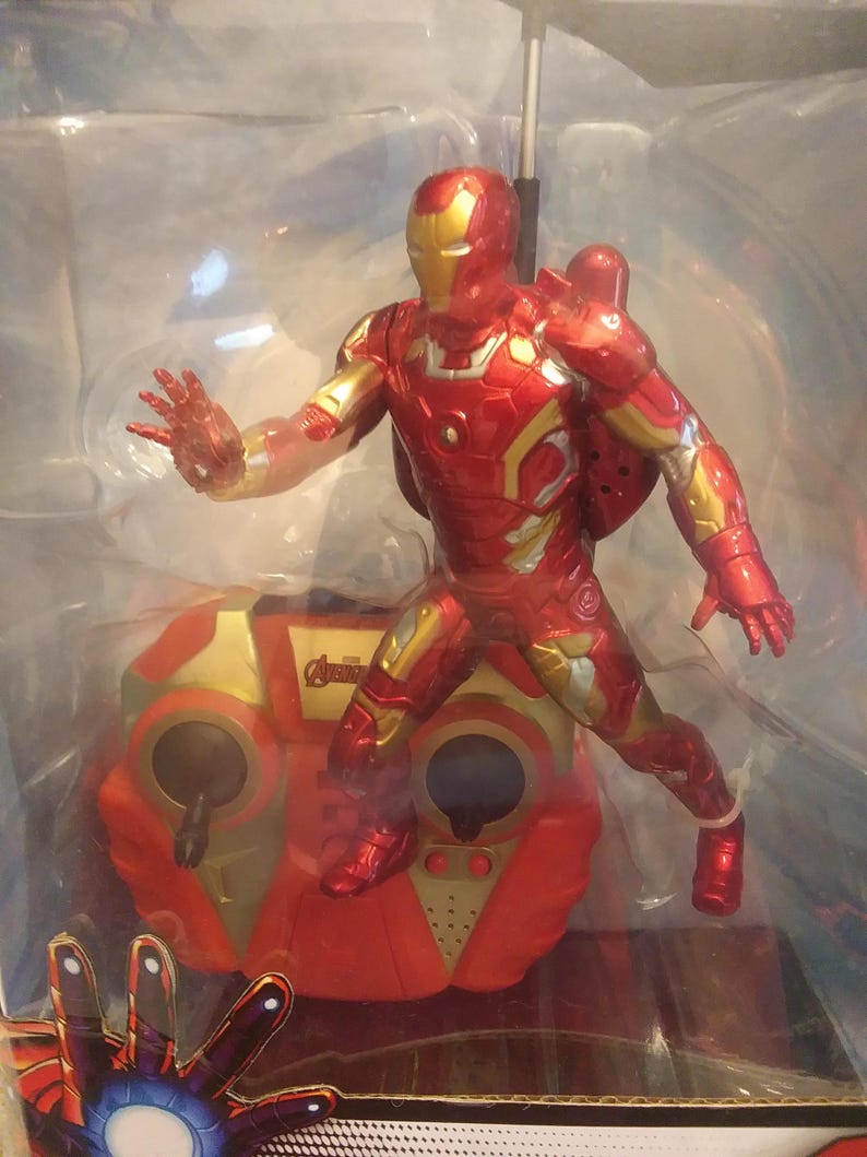 May include: A red and gold remote control toy of Iron Man flying with a red and gold remote control. The remote control has the Avengers logo on it.
