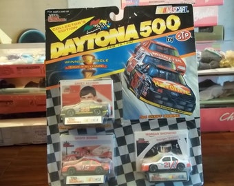 May include: A collector's edition set of three NASCAR die-cast cars from the 1992 Daytona 500. The cars are in a clear plastic package with a red, white, and blue checkered background. The cars are of Davey Allison, Geoff Bodine, and Morgan Shepherd. The package also includes a card with a picture of each driver and their car.