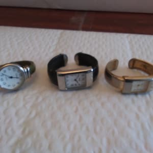 May include: Three silver wristwatches with different colored bands. The first watch has a green band, the second has a black band, and the third has a gold band.