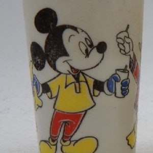 May include: White ceramic cup featuring a cartoon image of Mickey Mouse wearing a yellow shirt and red trousers. Mickey is holding a glass of liquid and smiling. The cup has the text "Walt Disney Productions" at the bottom.