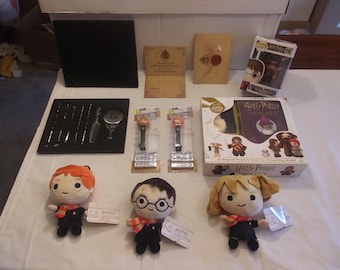 May include: A collection of Harry Potter themed items including plush toys, a book, a pop figure, and a set of tools. The plush toys are of Harry Potter, Ron Weasley, and Hermione Granger. The book is titled "Harry Potter: A History of Magic". The pop figure is of Harry Potter. The tools are in a black case and include a comb, a wand, and a mirror.