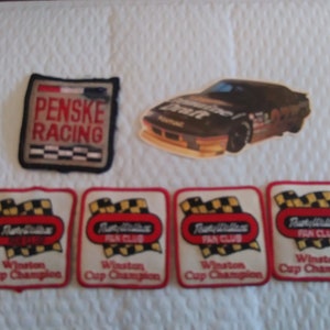 May include: Four white and red embroidered patches with the text "Rusty Wallace Fan Club Winston Cup Champion" and a checkered flag design. A black and white image of a race car with the text "Penske Racing" on a black and white patch.