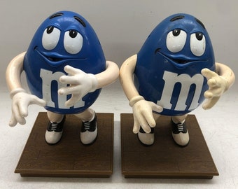 以下が含まれることがあります： 白い手袋と靴を履いた青いM&Mのフィギュア2体が、木製の台の上で踊っています。M&Mは白い目と笑顔をしていて、胸には「M」の文字が印刷されています。