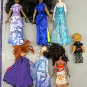 Puede incluir: Una colecci&oacute;n de ocho mu&ntilde;ecas, incluyendo princesas, una reina y un pr&iacute;ncipe. Las mu&ntilde;ecas llevan todas diferentes atuendos, incluyendo un vestido azul, un vestido morado, un vestido blanco, un vestido rojo, un vestido amarillo, un vestido verde, un vestido marr&oacute;n y un vestido negro.