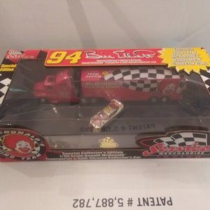 May include: A red 1/64 scale die-cast toy truck with a white checkered roof and "McDonald's Racing Team" printed on the side. The truck is carrying a smaller red die-cast car with the number 94 and "Bill Elliott" printed on the side. The box is labeled "Special Collector's Edition" and "Patent # 5,887,782".