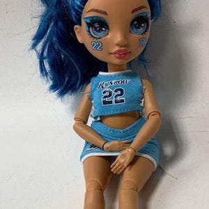May include: A blue and white cheerleading outfit with the word "Rainbow" and the number "22" on the front. The doll has blue hair with a white and blue bow, blue eyeshadow, and a pink mouth. The doll is wearing blue and white sneakers.