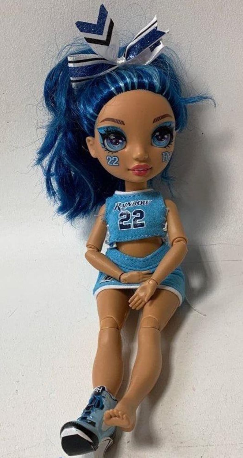 May include: A blue and white cheerleading outfit with the word "Rainbow" and the number "22" on the front. The doll has blue hair with a white and blue bow, blue eyeshadow, and a pink mouth. The doll is wearing blue and white sneakers.