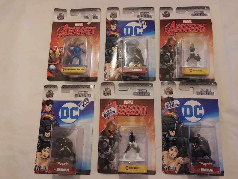 May include: Six packaged Marvel Avengers and DC Comics figurines. The figurines are in clear plastic cases attached to cardboard backings. The packaging features the Marvel Avengers and DC Comics logos, along with character names like Batman and Nick Fury.