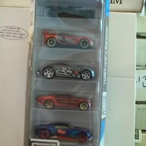 Op de afbeelding: Een vijf-pack Hot Wheels Justice League-speelgoedauto's in een doorzichtige plastic displaydoos. De auto's zijn rood, blauw, grijs en zwart met Justice League-personage-logo's. De doos heeft een Justice League-logo en de woorden "Justice League" erop gedrukt.