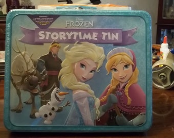 May include: A teal metal lunchbox featuring characters from Disney's Frozen. The box has the words "Frozen Storytime Tin" on the front, along with images of Elsa, Anna, Olaf, Kristoff, and Sven. The box is rectangular with a handle.