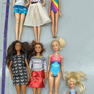 Puede incluir: Siete mu&ntilde;ecas Barbie con diferentes atuendos, que incluyen una blusa blanca con una falda azul, un vestido de lunares blanco y negro, una camisa a rayas con una falda roja, un traje de ba&ntilde;o azul, un traje de ba&ntilde;o arco&iacute;ris, un traje de ba&ntilde;o rosa y azul y un traje de ba&ntilde;o azul. Las mu&ntilde;ecas tienen diferentes colores y estilos de cabello.