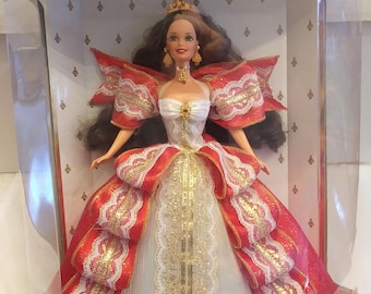 May include: A Barbie doll wearing a white gown with red and gold ribbon accents. The doll is in a clear plastic display box with a red and gold background. The box says "Happy Holidays" and "Special Edition Barbie".