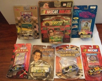 Op de afbeelding: Een verzameling NASCAR-modelwagens in verschillende verpakkingen. De auto's zijn in verschillende kleuren en met verschillende coureurs en sponsors. De verpakkingen bevatten de woorden "NASCAR Rules!" en "Collector's Series".