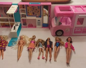 May include: A pink Barbie camper van and playset with several Barbie dolls. The camper has a white stripe and black wheels. The playset includes a closet, shelves, and a dressing area. The dolls are wearing various outfits.