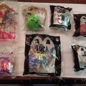May include: A collection of six clear plastic bags containing various toys and characters. The bags are labeled with the names of the characters, including "Luca", "Pons Gone Wrong", and "Gamer B-Bot".