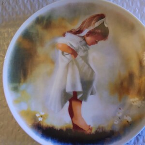 May include: A white porcelain plate with a painted image of a young girl in a white dress with a red bow in her hair. The girl is looking down at her feet, which are bare. The background is a soft, blurred watercolor of yellow, green, and blue.